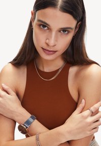Bruine mouwloze top, zilveren ketting, rosé gouden horloge met een donkere wijzerplaat, bijpassende kettingarmband en ring op gekruiste armen.