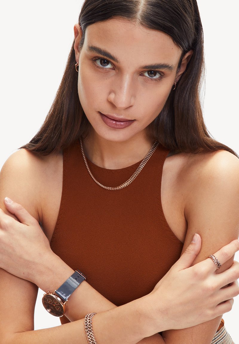Bruine mouwloze top, zilveren ketting, rosé gouden horloge met een donkere wijzerplaat, bijpassende kettingarmband en ring op gekruiste armen.