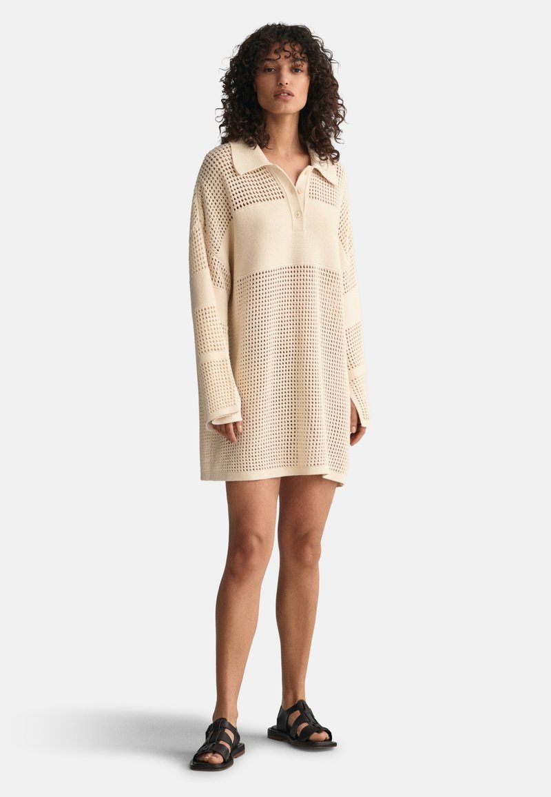 Sweterkowa sukienka w kolorze kremowym z kołnierzykiem, luźny fason, tekstura siateczkowa i długie rękawy. Na nagich nogach noszone są czarne sandały na paski.