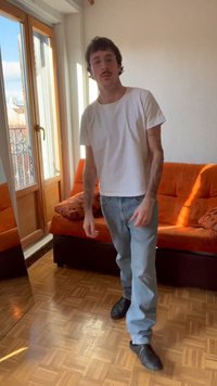 Hombre con bigote, gafas, camiseta blanca y vaqueros, de pie en una habitación iluminada por el sol con un sofá naranja y suelo de madera cerca de una ventana.