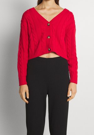 Femme portant un cardigan court rouge à torsades avec trois boutons et un pantalon noir taille haute, debout devant un fond uni.