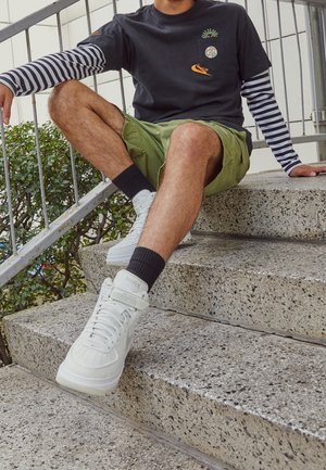 Zapatillas altas blancas, pantalones cortos verde olivo, camiseta negra con un gráfico de piña y mangas largas a rayas, sentado en escalones de piedra.