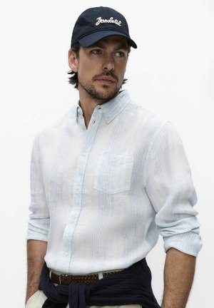 Man in een lichtblauw gestreept overhemd met opgerolde mouwen, een donkerblauwe pet en een donkere trui om zijn middel gestrikt, kijkt opzij.