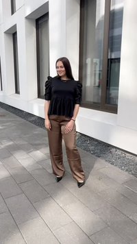 Černý peplum top s nafouknutými rukávy, v kombinaci s hnědými saténovými kalhotami s širokými nohavicemi a černými podpatky; hladké textury a čisté linie.