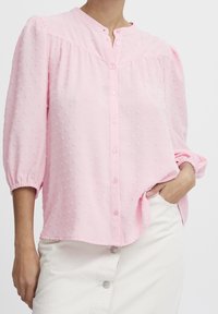Blusa rosa chiaro con chiusura a bottoni, collo rotondo e maniche a sbuffo. Il tessuto testurizzato presenta piccole macchie rialzate per dettagli.