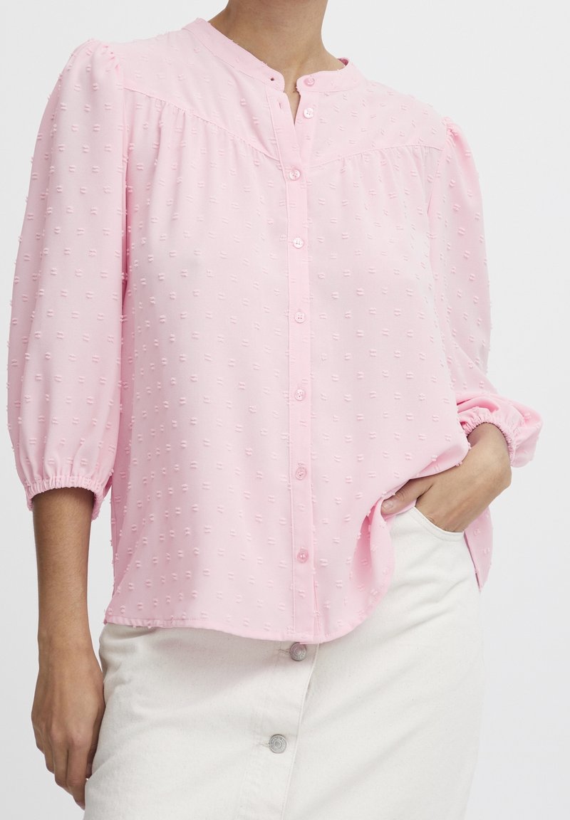 Blusa rosa chiaro con chiusura a bottoni, collo rotondo e maniche a sbuffo. Il tessuto testurizzato presenta piccole macchie rialzate per dettagli.