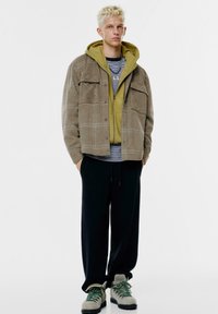 Ruskea villakangastakki vihreän hupparin päällä, kerroksittain raidallisen paidan kanssa. Mustat verkkarit ja harmaa-vihreät lenkkarit mustilla pohjilla.