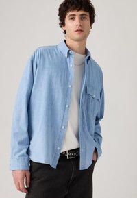 Lyseblå skjorte med knapper i blød stof, med en brystlomme, parret med en hvid T-shirt og sorte bukser med et nitterbælte.