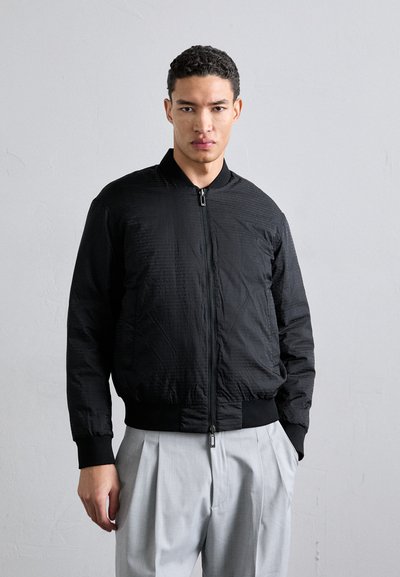 Sort bomberjakke med lynlås foran, ribbede manchetter og kant, med en struktureret overflade og en let tætsiddende pasform. Sammen med lysegrå bukser.