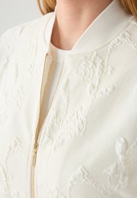 Witte bomberjack met een gestructureerd bloemenpatroon, geribbelde kraag en gouden ritsdetails. Zachte stof met een gladde afwerking.