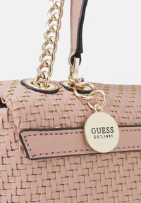 Gewebte pinke Handtasche mit goldfarbener Hardware, ausgestattet mit einer Kettenriemen und einem runden Markenanhänger mit der eingeprägten Aufschrift "GUESS EST. 1981".