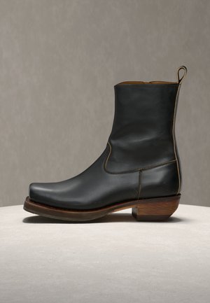 Μαύρο δερμάτινο μποτάκι μέχρι τον αστράγαλο με τετράγωνη μύτη, ξύλινο τακούνι και ταινία έλξης, σε ουδέτερη επιφάνεια και φόντο.