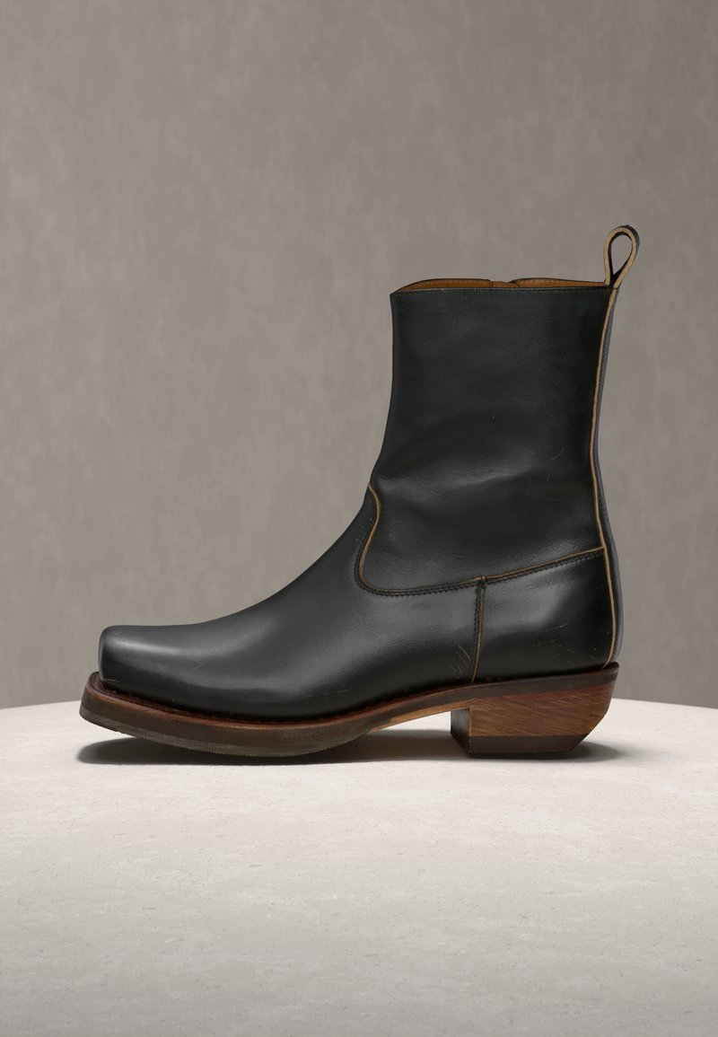 Μαύρο δερμάτινο μποτάκι μέχρι τον αστράγαλο με τετράγωνη μύτη, ξύλινο τακούνι και ταινία έλξης, σε ουδέτερη επιφάνεια και φόντο.