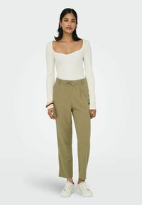 Le modèle se tient sur un fond neutre, portant un haut à manches longues blanc et un pantalon vert olive, associé à des baskets blanches.