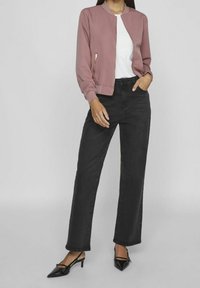 Woman wearing mauve bomber jacket, white t-shirt, black high-waist jeans, and black pointed-toe heels, standing with one hand in pocket.