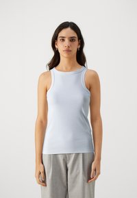Haut sans manches bleu clair en tissu lisse, avec un col montant et une coupe ajustée. Porté avec un pantalon gris à motifs.