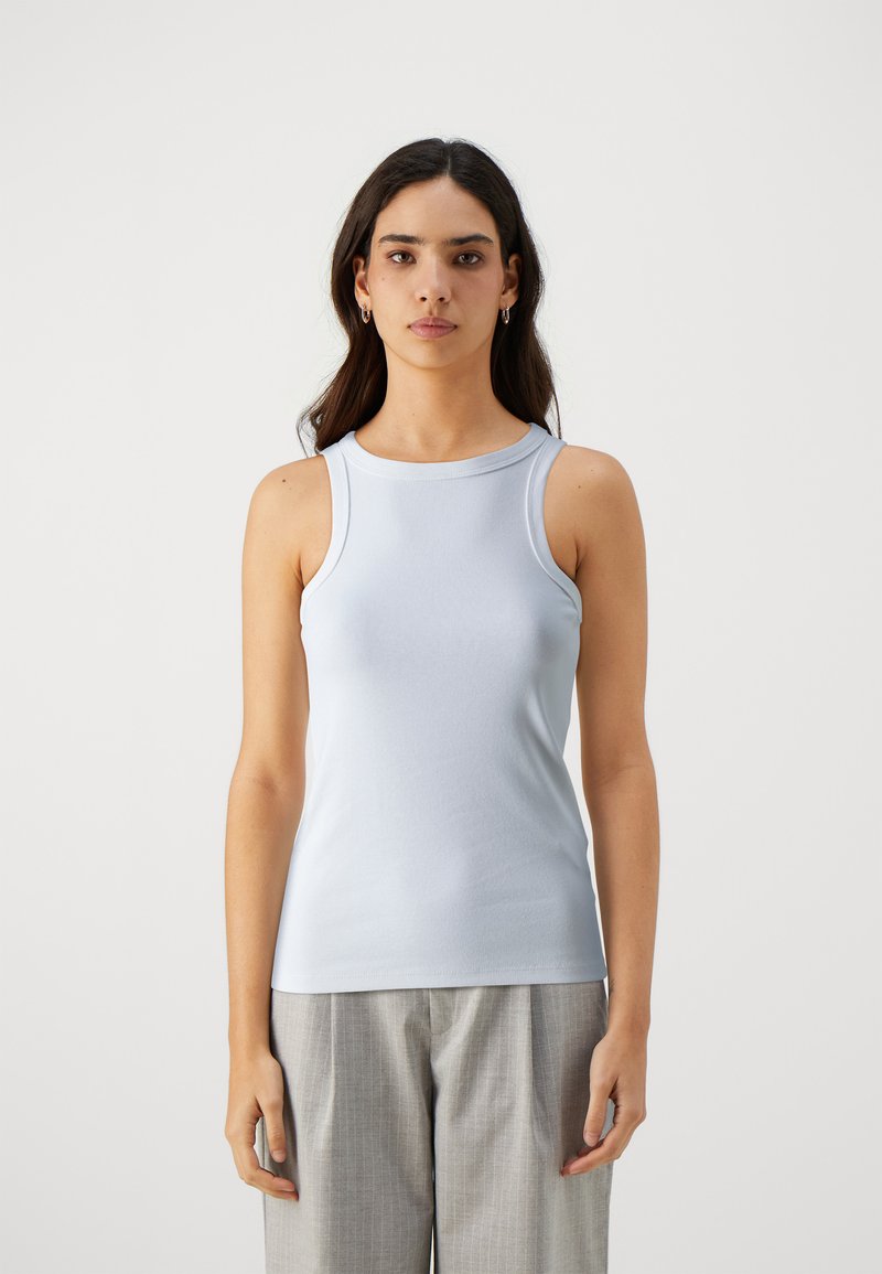 Haut sans manches bleu clair en tissu lisse, avec un col montant et une coupe ajustée. Porté avec un pantalon gris à motifs.