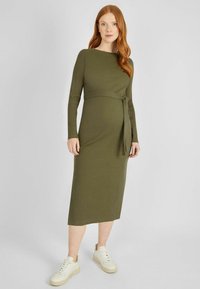 Olivegrünes, geripptes Midikleid mit langen Ärmeln und einem Bindegürtel in der Taille, kombiniert mit weißen Sneakers und einer glatten Textur.