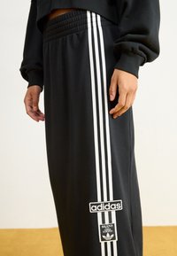Fekete sportnadrág, három fehér csíkkal az oldalán. Rugalmas derékpánttal rendelkezik, és adidas logóval ellátott a lábszárán.
