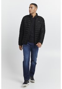 Black puffer jacket with horizontal quilted pattern, high collar, and zip closure. Worn over a black shirt and paired with blue jeans and black sneakers.