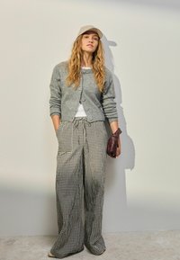 Grijze wollen cardigan met knopen, witte top eronder, losse zwart-wit geblokte broek, houdt een bordeauxrode handtas vast, staat tegen een muur.