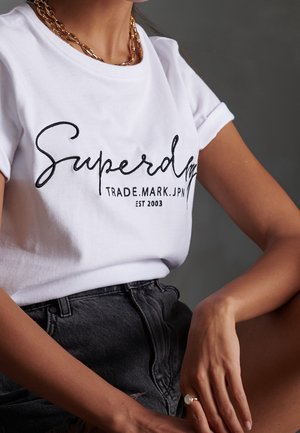Personne portant un t-shirt blanc Superdry, un short en jean noir, un collier chaîne en or et une bague, assise avec un bras reposant sur le genou, sur fond gris.
