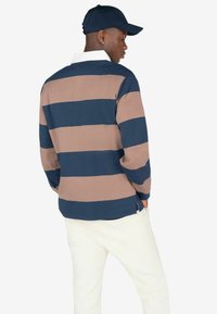 Polo a maniche lunghe a righe blu navy e beige con colletto bianco, indossato con pantaloni color crema. Cappellino in materiale blu navy abbinato.