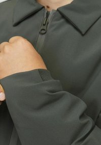 Tamsiai žalia striukė su struktūrizuota apykakle, ilga užtrauktuku ir elastinėmis rankogaliais. Lygus paviršius su subtiliu blizgesiu.
