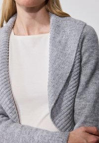 Szürke kötött kardigán bordázott textúrával és sálgallérral, fehér felsőn viselve. Puha anyag, szűk szabású ujjak, és nyitott elülső kialakítás.