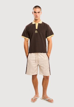 Męski model stojący z rękami w kieszeniach, ubrany w brązowe polo z żółtą lamówką, beżowe szorty z troczkiem i białe klapki.