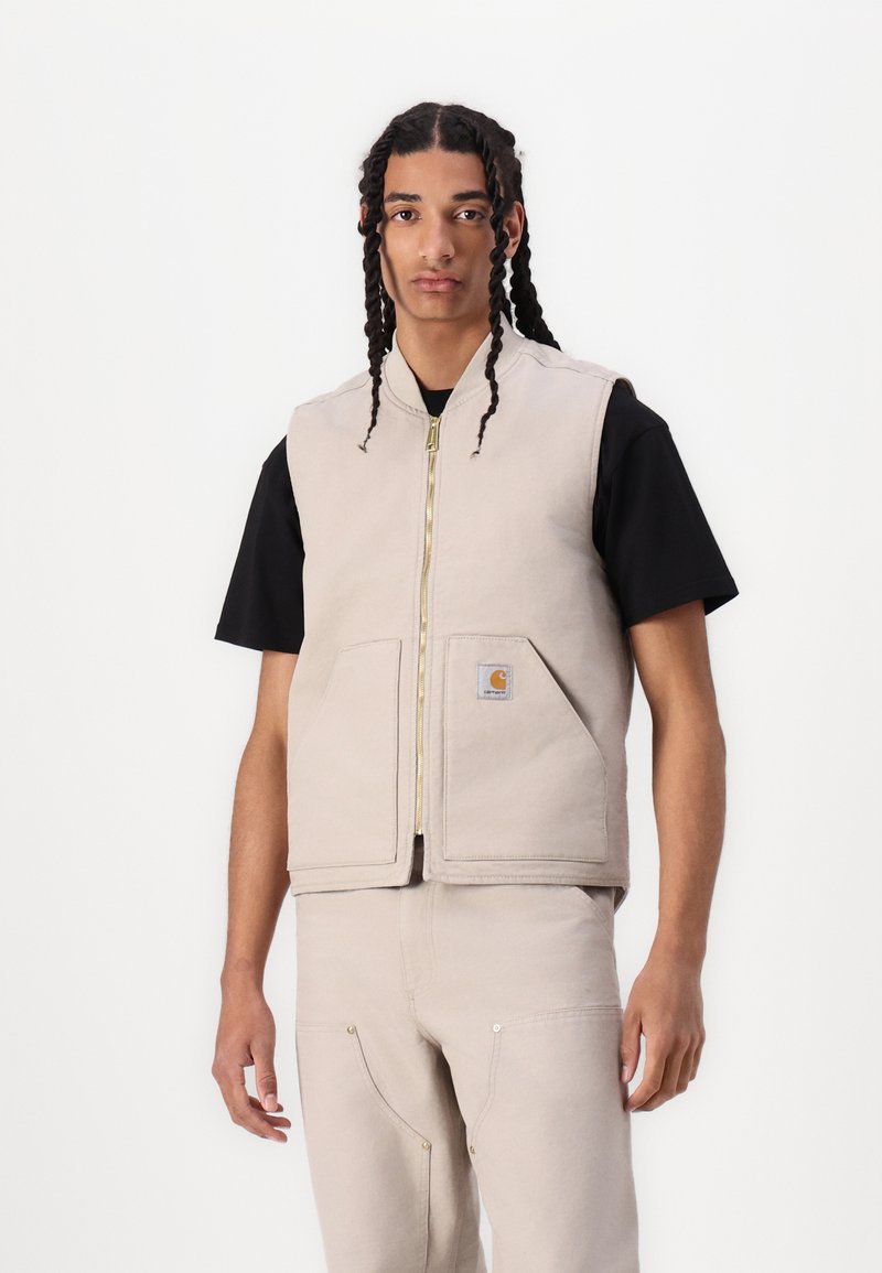 Beige bomullsvest med glidelås foran, rund halslinje og to frontlommer. Båret over en svart t-skjorte, med et synlig merke.
