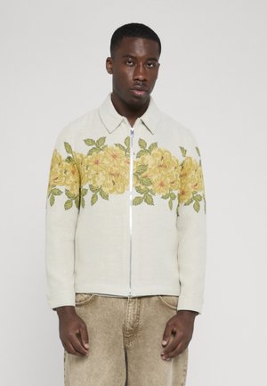 Mand iført en let jakke med et gult blomsterprint over brystet og de øverste ærmer, kombineret med beige bukser.