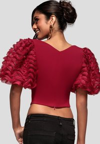 Kobieta ubrana w dopasowaną czerwoną bluzkę z krótkimi, warstwowymi falbaniastymi rękawami, w połączeniu z czarnymi spodniami, uśmiechająca się i zwrócona bokiem.