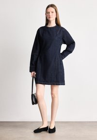 Σκούρο τζιν φόρεμα με άνετη γραμμή, στρογγυλή λαιμόκοψη και μακριά μανίκια. Διαθέτει διακριτές ραφές και πλαϊνές τσέπες. Φορεμένο με μαύρες μπαλαρίνες.