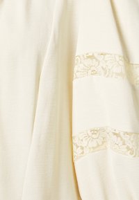 Tissu couleur crème avec un tissage texturé, présentant des accents en dentelle dans un motif floral le long d'un bord. Matériau doux et léger.