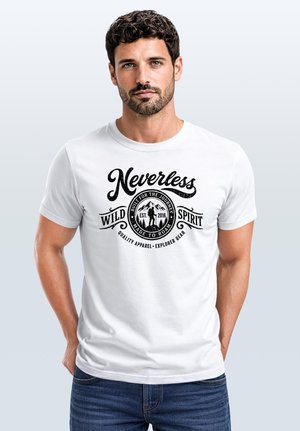 Mann mit kurzen dunklen lockigen Haaren und Bart, der ein weißes T-Shirt mit schwarzem Text "Nevertheless Wild Spirit" und Berggrafik trägt, Hände in den Taschen.