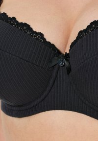 Zwarte geribbelde beha met kantafwerking aan de randen, met een centrale strikaccent en een subtiele knoopdetail. Glad stoftextuur.