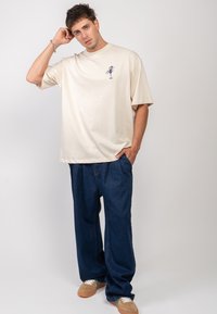 Krémové oversized tričko s modrou grafikou, v kombinaci s volnými tmavě modrými džíny. Model má na sobě krémové a modré tenisky.