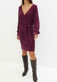 Robe portefeuille en maille couleur bourgogne foncé, avec un col en V, des manches longues, une ceinture nouée à la taille, et une jupe ajustée. Associée à des bottes hautes jusqu’au genou.