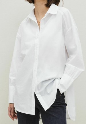 Woman wearing loose white button-up shirt with long sleeves tucked partially into dark jeans, hand in pocket, neutral background.