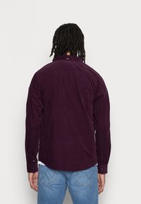 Camicia in velluto a coste bordeaux con maniche lunghe, polsini con bottone e colletto classico. Presenta una texture sottile e un orlo curvo.