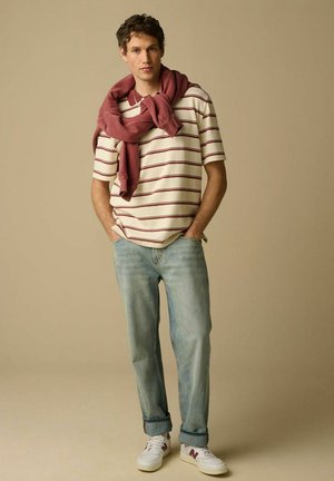 Homme portant un T-shirt crème à rayures rouges, un jean bleu clair, des baskets blanches, avec un sweat-shirt bordeaux drapé sur ses épaules.