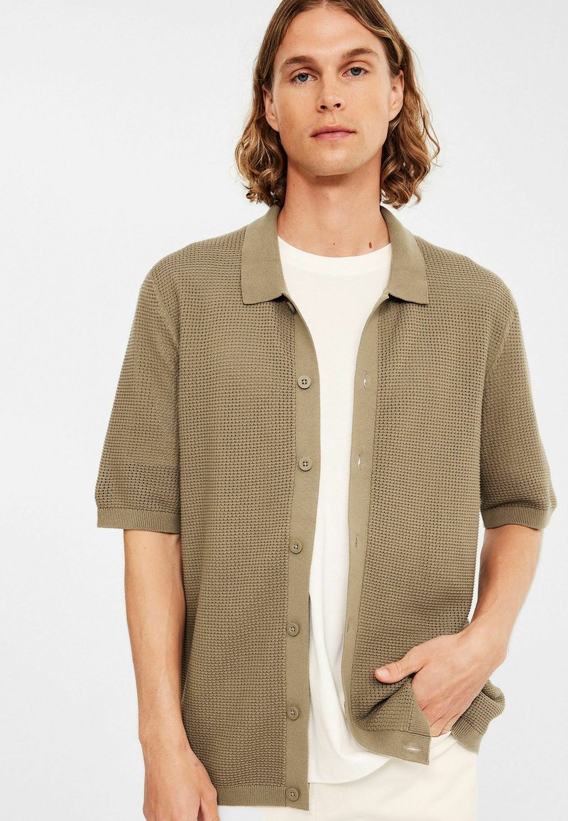 Jonge man met schouderlang haar, gekleed in een beige knoopshirt met korte mouwen over een wit t-shirt, met één hand in zijn zak.