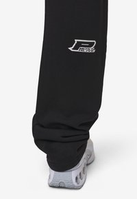 Svarta sweatpants med en tight mudd vid ankeln har en vit logotyp på benet. Kombinerat med en vit sneaker med grå accenter.