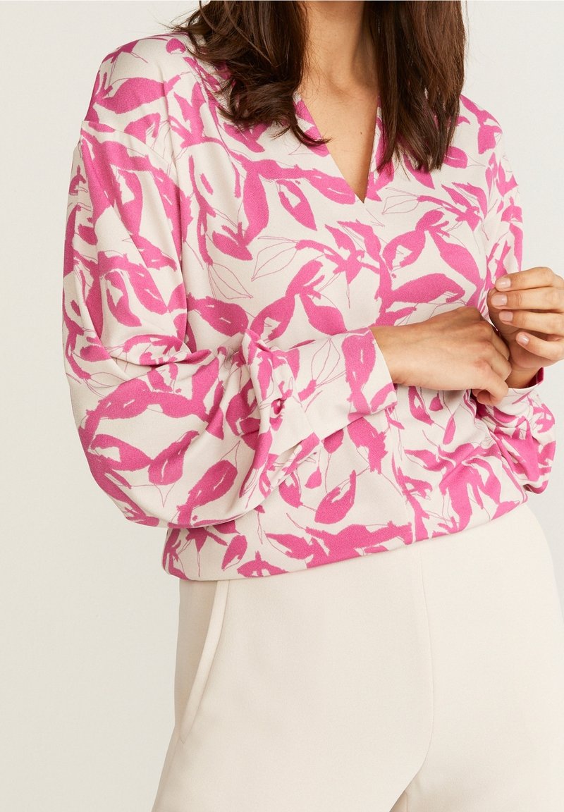 Femme portant un haut à manches longues rose et blanc avec des motifs abstraits de feuilles et un pantalon clair, mains doucement jointes.