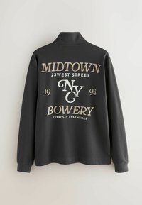 Czarna szara koszula z długim rękawem i kołnierzykiem. Na przodzie znajduje się biało-złoty napis: „MIDTOWN 23 WEST STREET NYC 1994 BOWERY EVERYDAY ESSENTIALS.”