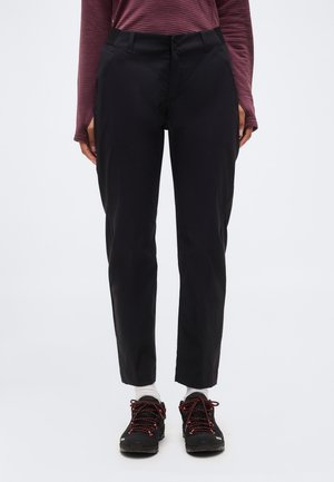 Osoba oblečená v černých kalhotách s rovnými nohavicemi, bílých ponožkách, černých turistických botách s červenými tkaničkami a dlouhém rukávu bordó trička.