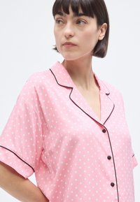 Ροζ πυτζάμα με κοντά μανίκια και λευκές πουά, μαύρη λεπτομέρεια κατά μήκος του γιακά και της λωρίδας με κουμπιά, με άνετη εφαρμογή.