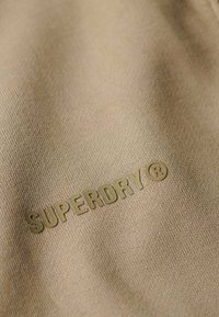 Tessuto beige con una texture liscia, caratterizzato da un logo "SUPERDRY ®" in rilievo di colore verde oliva. Il design è semplice e minimalista.