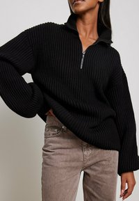 Schwarzer gerippter Pullover mit hohem Kragen und halber Reißverschluss, mit weitem Ärmel. Trägt helle, braune Strukturhosen.